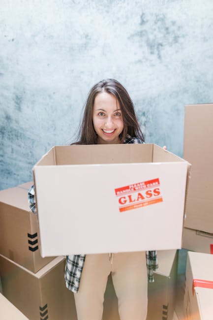 A woman with long brown hair smiling and looking at the camera while holding a large empty cardboard box marked 'FRAGILE GLASS' in red letters. She is standing indoors in front of a blue textured wall